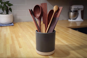 A dark gray ceramic utensil holder with a light natural wood base, filled with various wooden kitchen utensils including spoons and spatulas, set on a light wood countertop in a kitchen.