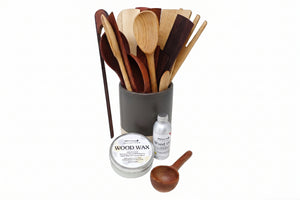 A set of wooden kitchen utensils in a dark gray holder, with a tin of wood wax, a small bottle of oil, and a wooden spoon in front, photographed on a white background.