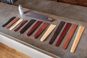 A set of wooden kitchen utensils, including spoons and a whisk, arranged on a gray stone countertop next to a small bottle of oil. The spoons and whisk feature various wood grain patterns in shades of dark brown, reddish-brown, beige, and gray.